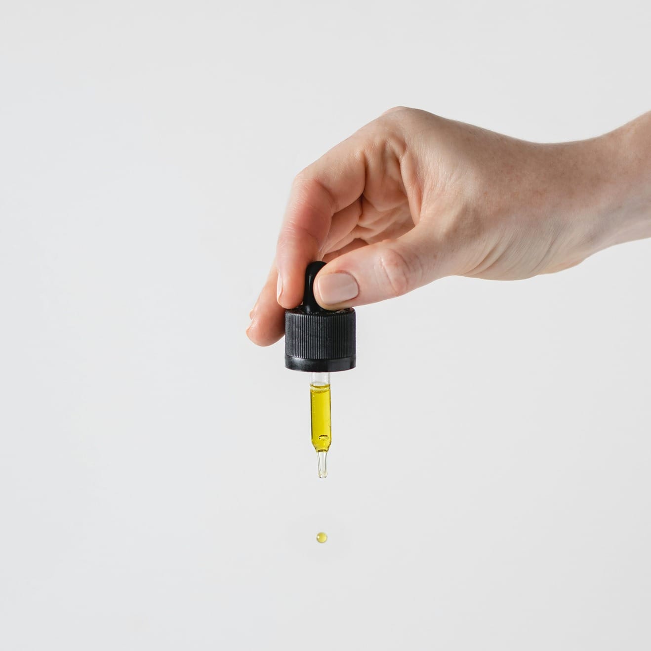 Hand hält eine Pipette mit CBD, aus der ein Tropfen CBD-Öl fällt, vor weißem Hintergrund